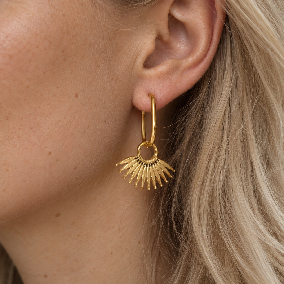 A close-up of a model's ear wearing a gold hoop earring with a dangling sunburst fan leaf charm.