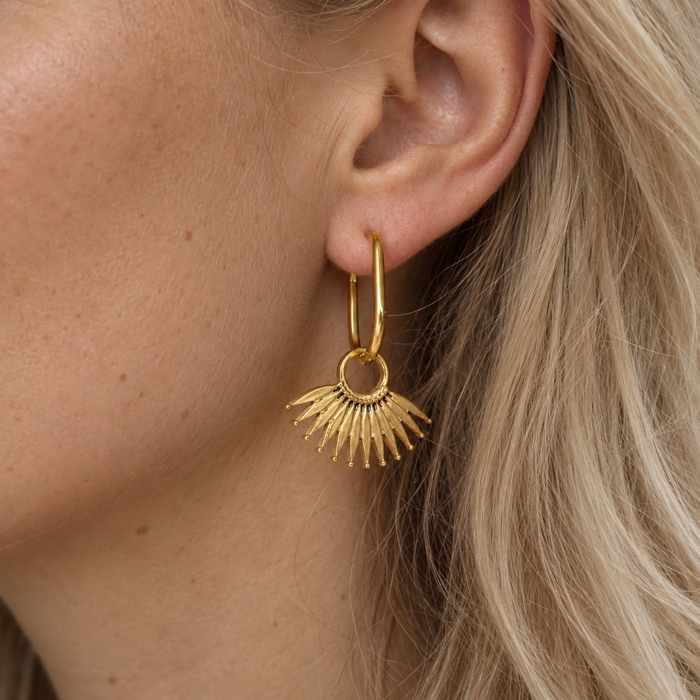A close-up of a model's ear wearing a gold hoop earring with a dangling sunburst fan leaf charm.