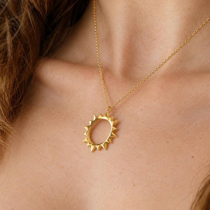 A gold pendant necklace featuring a circular sunburst design, worn on a model's neck.