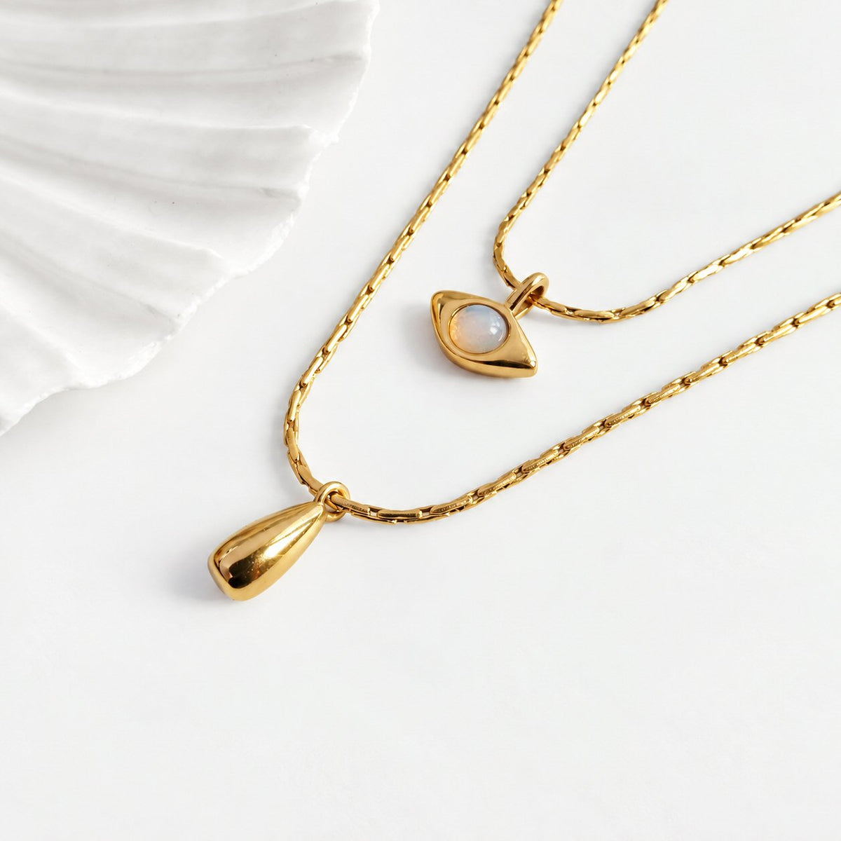 Two gold-tone necklaces displayed on a clean white surface. One necklace features a teardrop-shaped pendant, while the other has an eye-shaped pendant with a white moonstone center.
