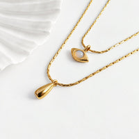 Two gold-tone necklaces displayed on a clean white surface. One necklace features a teardrop-shaped pendant, while the other has an eye-shaped pendant with a white moonstone center.