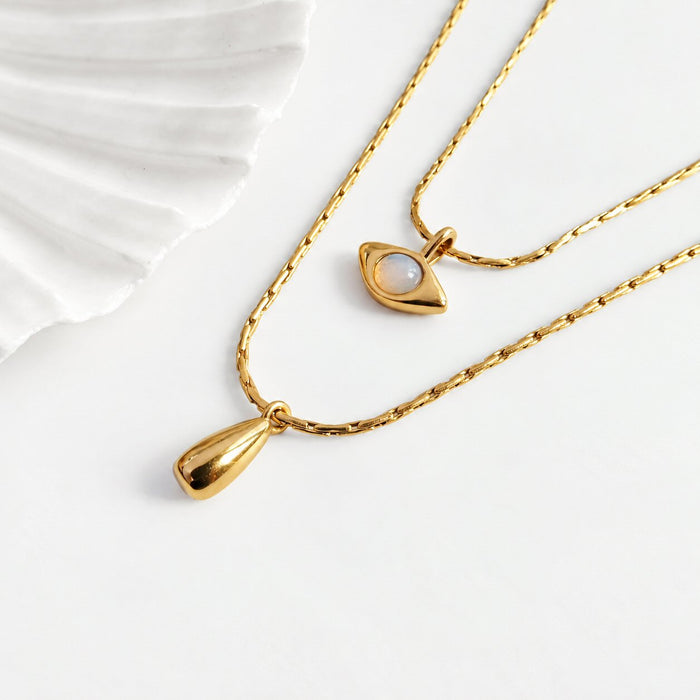 Two gold-tone necklaces displayed on a clean white surface. One necklace features a teardrop-shaped pendant, while the other has an eye-shaped pendant with a white moonstone center.