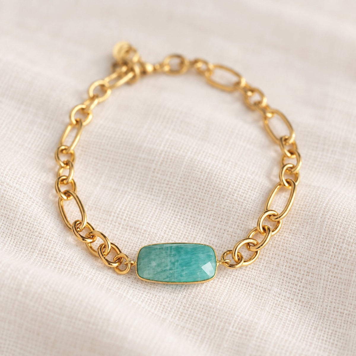 A gold chain bracelet featuring a rectangular, bezel-set turquoise stone, displayed on a textured off-white linen fabric background.