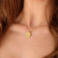 A close-up of a model's neck and chest wearing a delicate gold chain necklace with a textured, circular coin-style pendant.