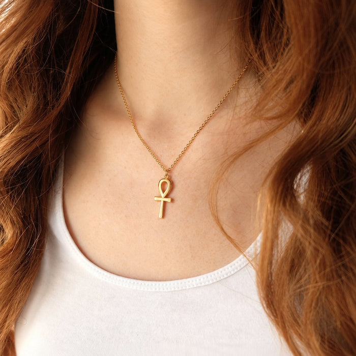 A close-up of a model wearing a delicate gold chain necklace with an ankh pendant, styled with a white ribbed tank top.