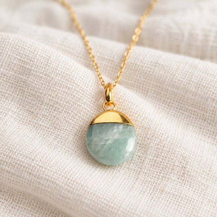 A gold chain necklace with a round pendant split horizontally into a polished gold top half and a light green stone bottom half, displayed on a textured cream linen fabric.