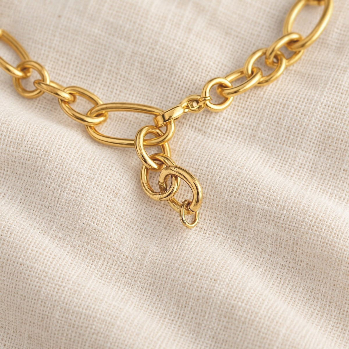 A close-up of a polished gold chain necklace with a lobster clasp and interlocking links, laid on a textured off-white linen fabric background.