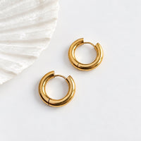 A pair of chunky gold huggie hoop earrings with a textured, ribbed finish, displayed on a clean white surface next to a white shell.