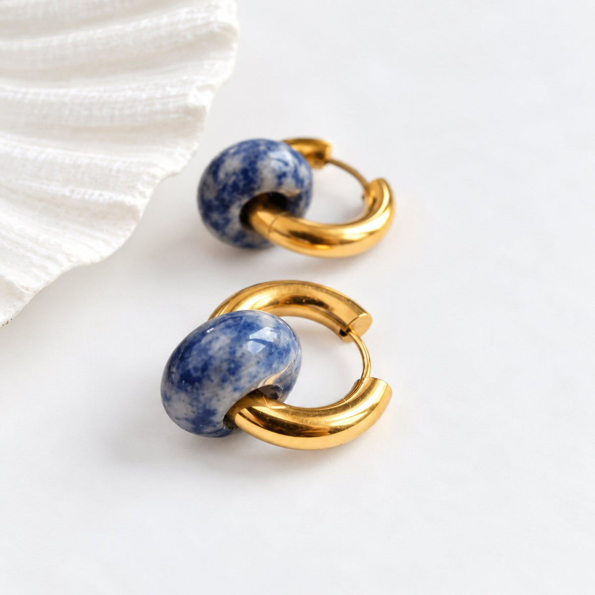 A pair of gold hoop earrings with polished blue and white speckled stone beads, displayed on a clean white surface next to a white textured shell.