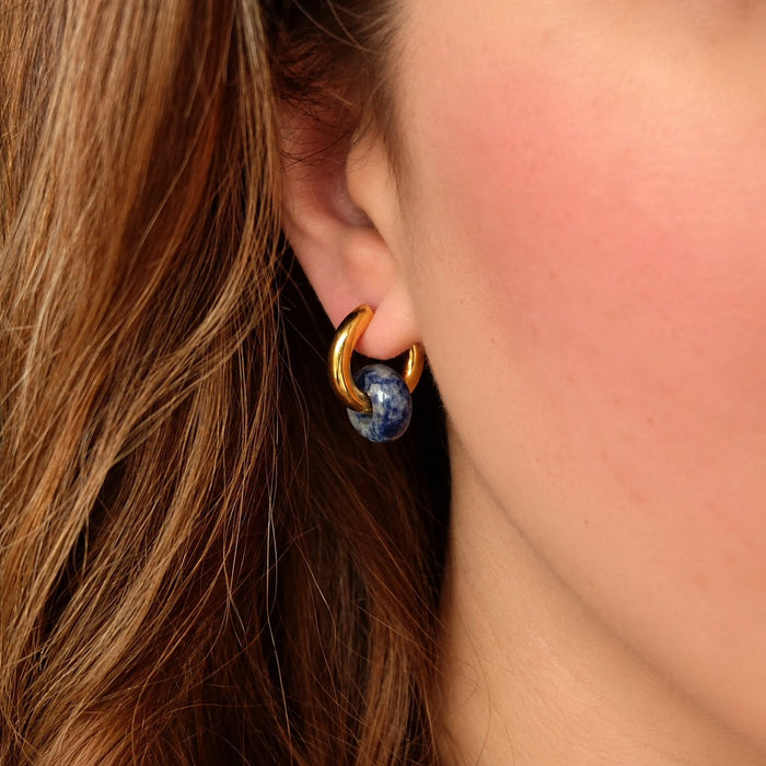 A close-up of a person's ear wearing a gold huggie hoop earring with a small round blue gemstone accent.