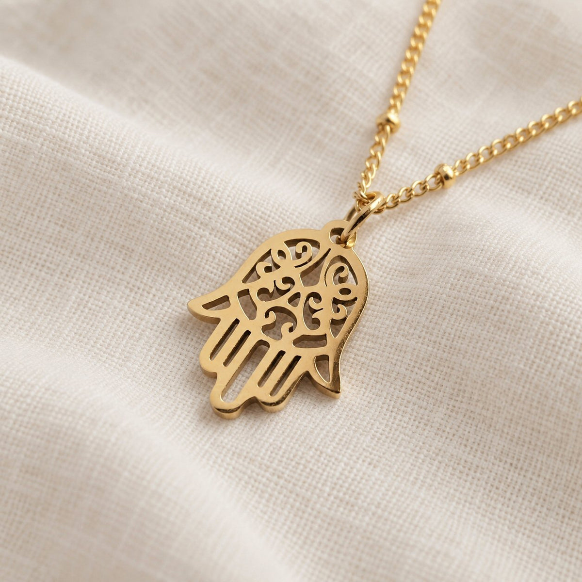 A gold-tone Hamsa hand pendant necklace with intricate cutout details, resting on a textured beige linen fabric background.