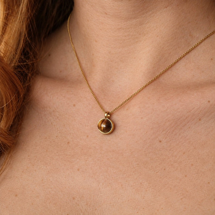 A close-up of a person's neck and chest wearing a delicate gold chain necklace with a small round bezel-set brown gemstone pendant.
