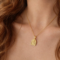 A close-up of a person wearing a delicate gold chain necklace with a small, detailed gold pendant featuring a religious figure, set against warm skin tones and brown hair.