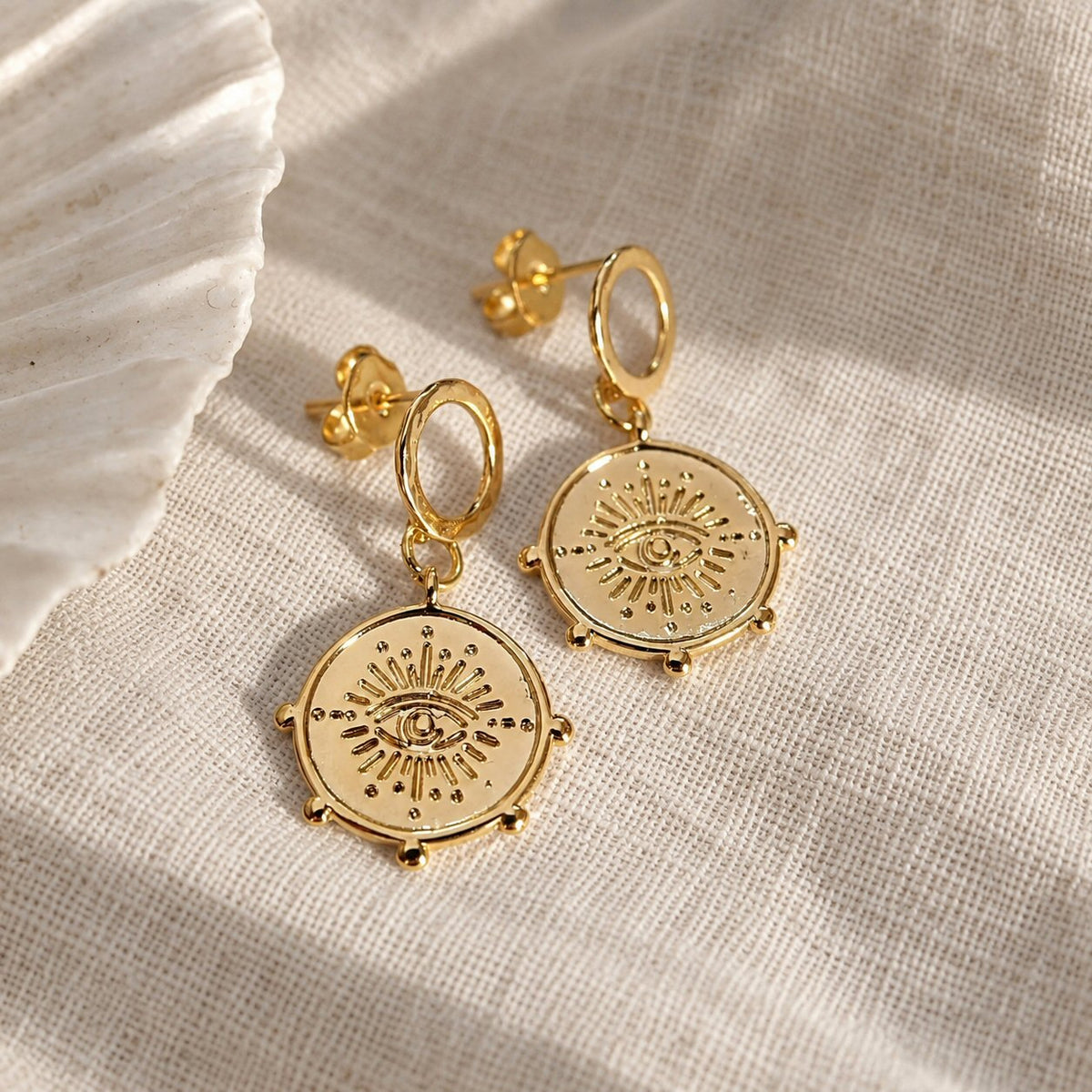 A pair of gold-toned dangle earrings with circular sunburst and eye motifs, displayed on a textured beige fabric next to a white shell.