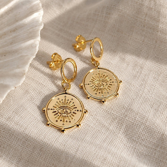 A pair of gold-toned dangle earrings with circular sunburst and eye motifs, displayed on a textured beige fabric next to a white shell.