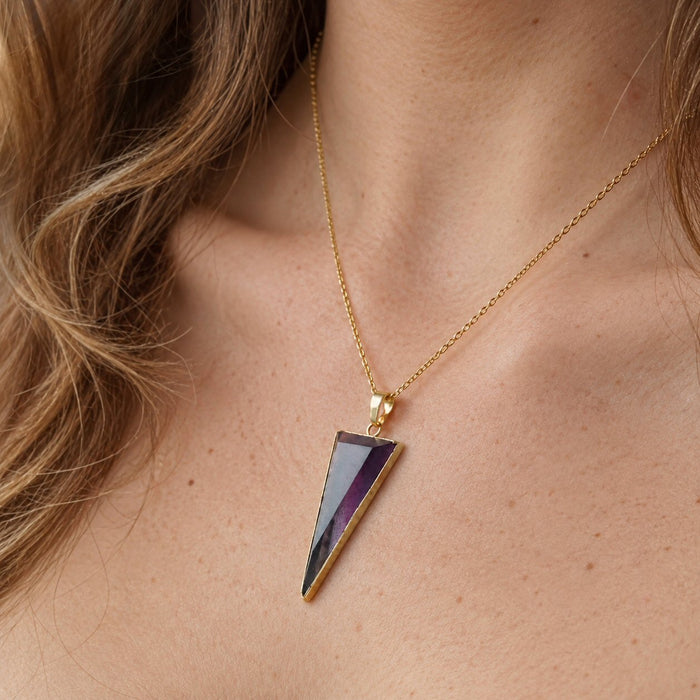a woman's neck and chest wearing a gold chain necklace with a triangular  pendant featuring a faceted purple and blue gemstone (fluorite) set in gold.