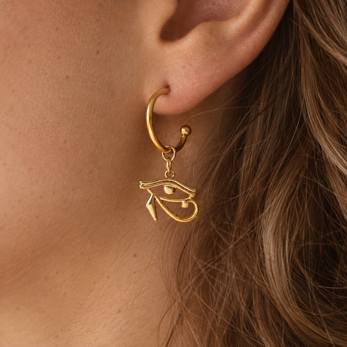 A close-up of a person's ear wearing a gold hoop earring with a dangling Egyptian eye charm.