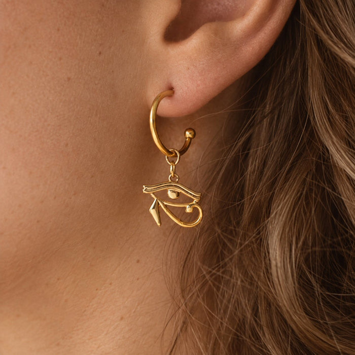 A close-up of a person's ear wearing a gold hoop earring with a dangling Egyptian eye charm.