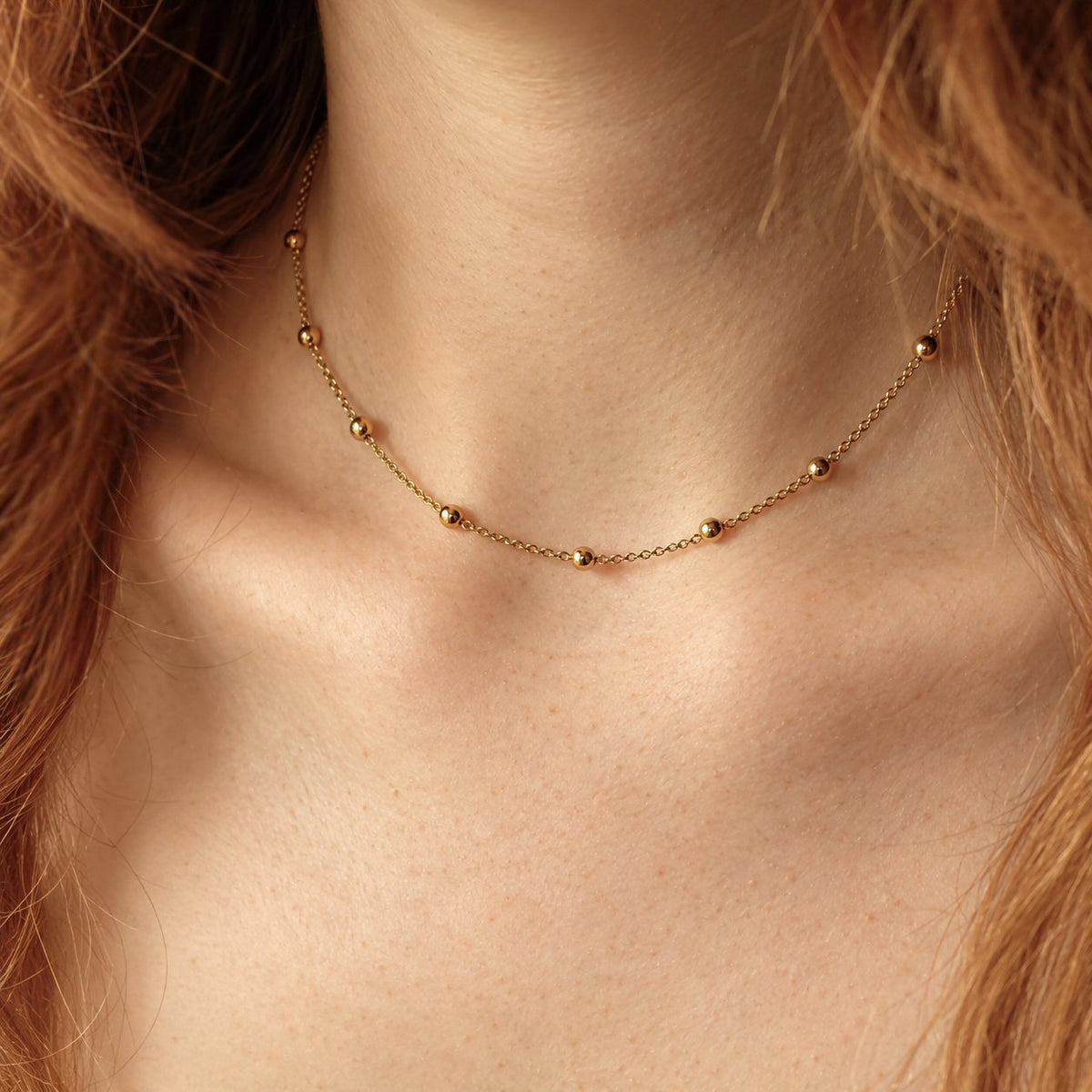 A delicate gold chain necklace with small spaced beads worn on a model's neck, shown in a close-up lifestyle shot.