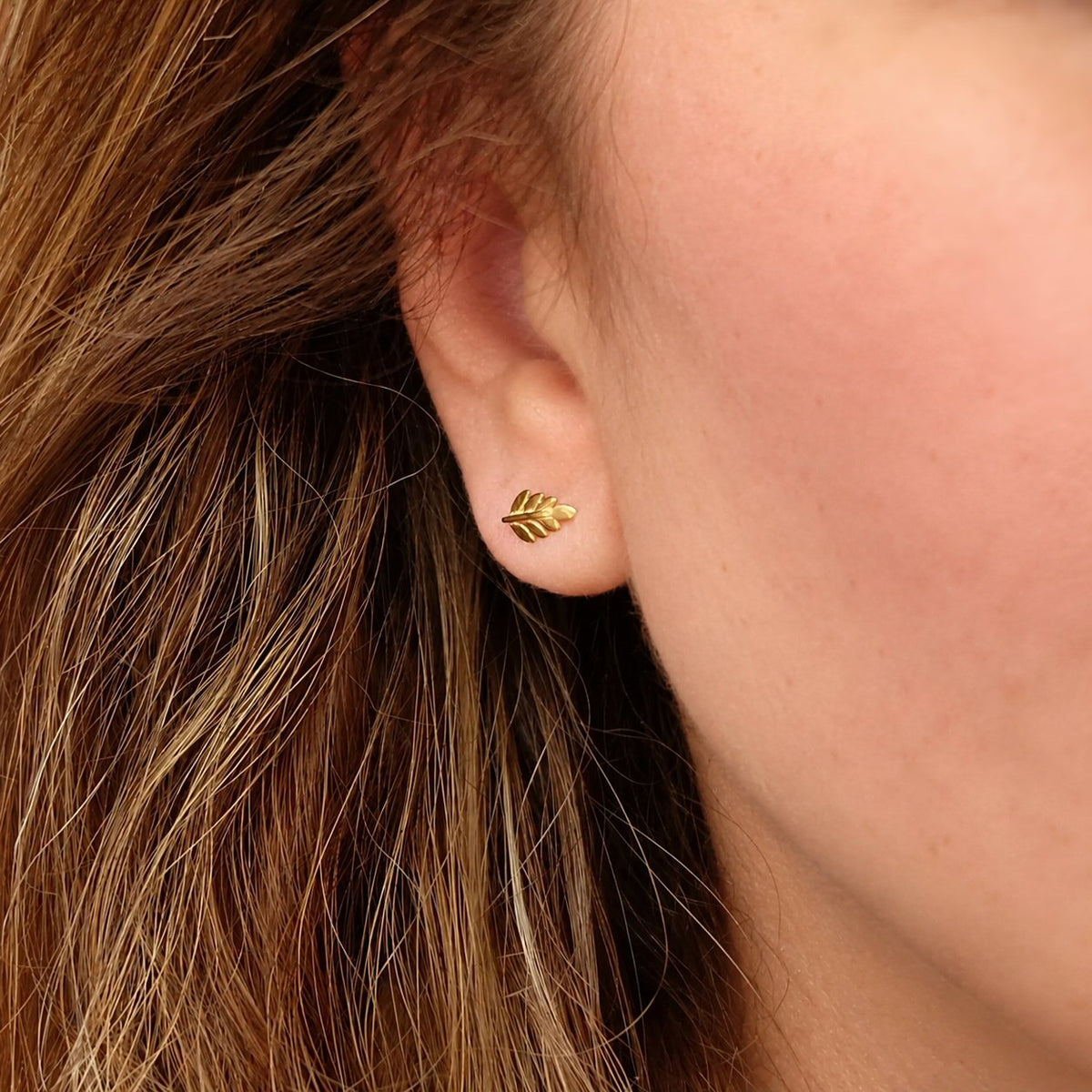 A close-up of a person's ear wearing a small gold leaf stud earring, with brown hair and skin visible.
