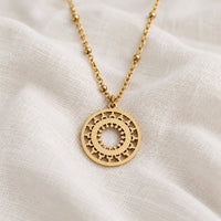 A gold-tone necklace with a circular cutout pendant featuring sunburst and star details, resting on draped white linen fabric.