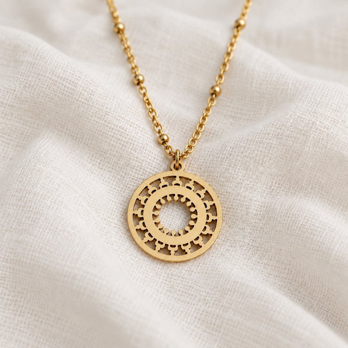 A gold-tone necklace with a circular cutout pendant featuring sunburst and star details, resting on draped white linen fabric.