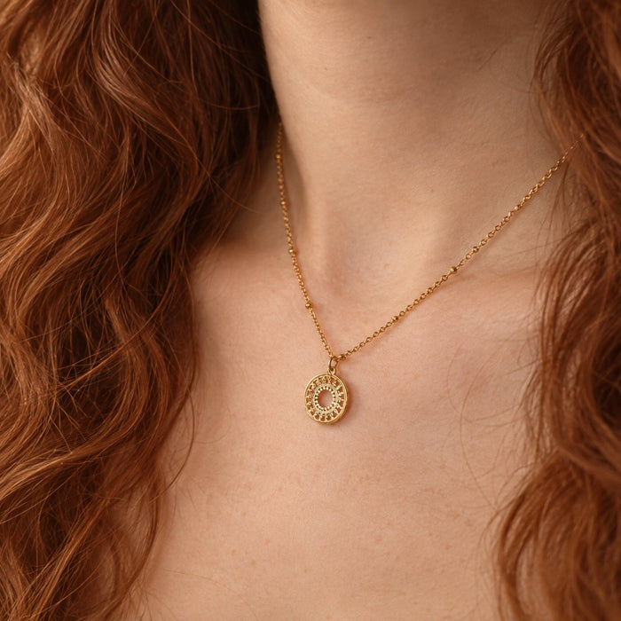 A close-up of a gold necklace with a circular sunburst pendant worn by a model with wavy red hair.