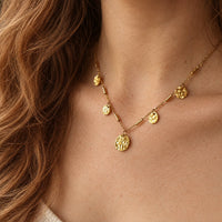 A close-up of a woman's neck and chest wearing a delicate gold necklace with multiple textured disc pendants and small cylindrical links.
