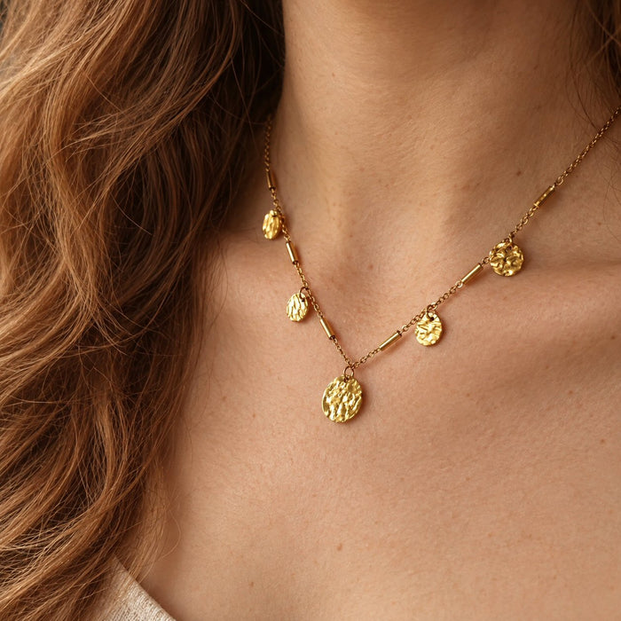 A close-up of a woman's neck and chest wearing a delicate gold necklace with multiple textured disc pendants and small cylindrical links.