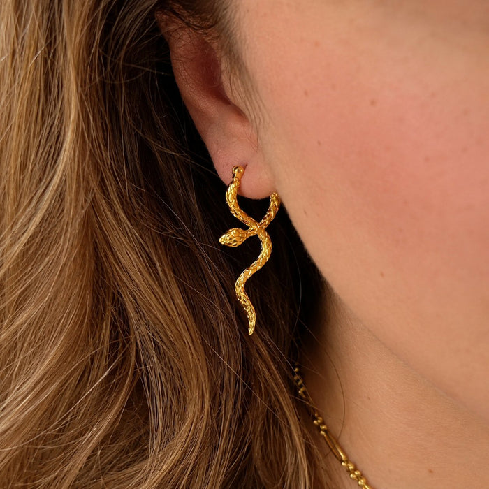 A close-up of a model's ear wearing a textured gold snake-shaped stud earring, with her hair and skin visible.
