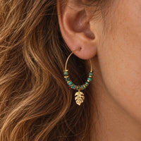 A close-up of a person's ear wearing a gold hoop earring adorned with small turquoise-blue beads and a dangling gold leaf charm.