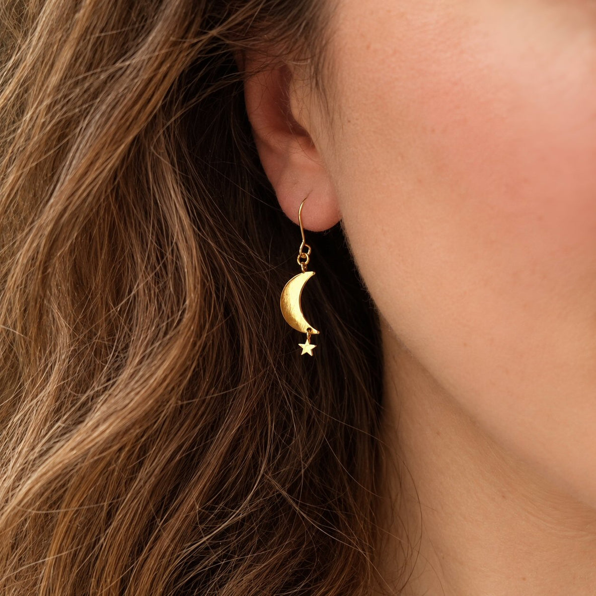 A close-up of a model's ear wearing a delicate gold crescent moon and star dangle earring, with her brown hair and skin visible.
