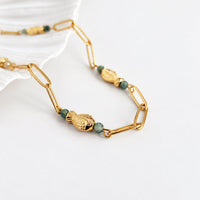 A gold link chain necklace featuring textured gold fish charms and small green faceted beads, displayed on a clean white surface.