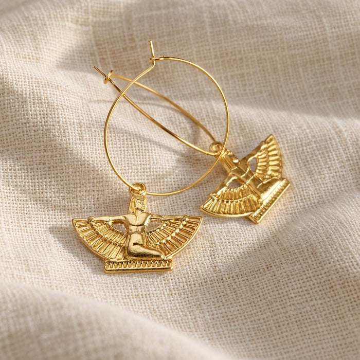A pair of gold hoop earrings with winged goddess charms, displayed on a textured beige linen fabric background.