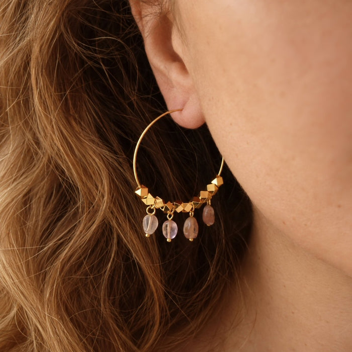 A close-up of a person's ear wearing a gold hoop earring adorned with faceted gold beads and four dangling rose quartz crystal drops.