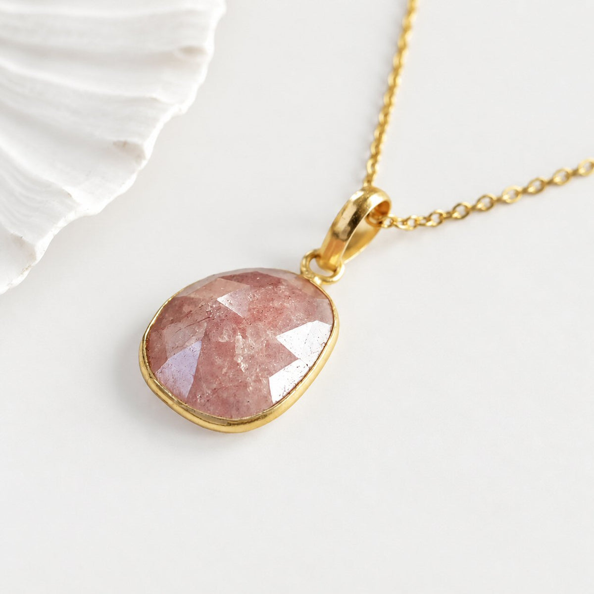 A gold pendant necklace featuring a faceted, teardrop-shaped pink gemstone, displayed on a clean white surface next to a white seashell.