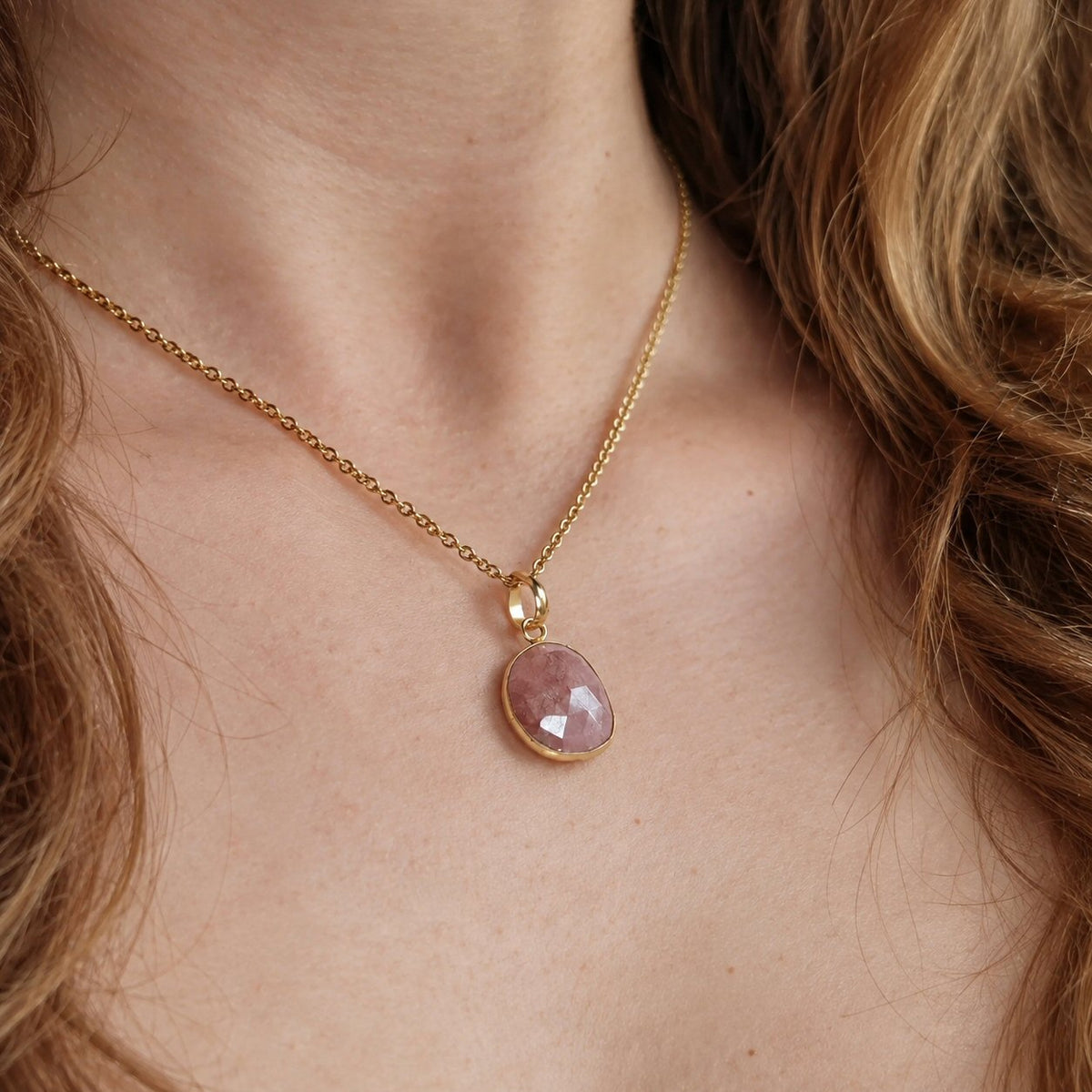 A close-up of a gold chain necklace with a faceted pink gemstone pendant, worn on a model's neck.