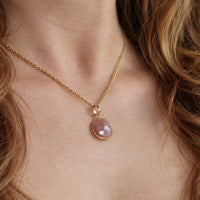 A close-up of a gold chain necklace with a faceted pink gemstone pendant, worn on a model's neck.