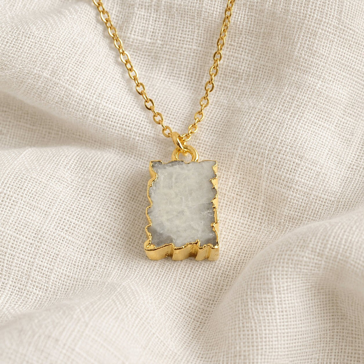 A gold chain necklace with a rectangular pendant featuring a pale grayish-white stone with natural jagged edges, displayed on a textured off-white linen fabric background.