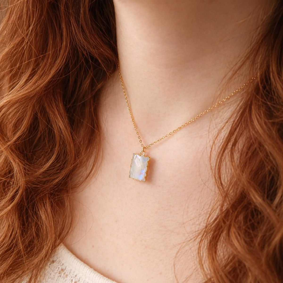 A close-up of a woman wearing a delicate gold chain necklace with a small, rectangular iridescent pendant on a warm skin tone with red hair.