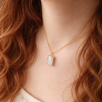 A close-up of a woman wearing a delicate gold chain necklace with a small, rectangular iridescent pendant on a warm skin tone with red hair.