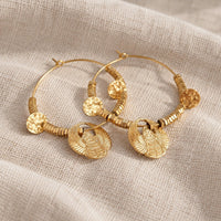 A pair of gold hoop earrings with textured circular beads and winged sun motifs, displayed on a beige linen fabric background.