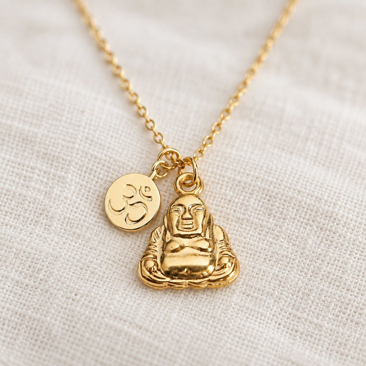 A gold-tone Buddha pendant necklace with a small Om symbol charm, displayed on a textured off-white linen fabric background.