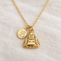 A gold-tone Buddha pendant necklace with a small Om symbol charm, displayed on a textured off-white linen fabric background.