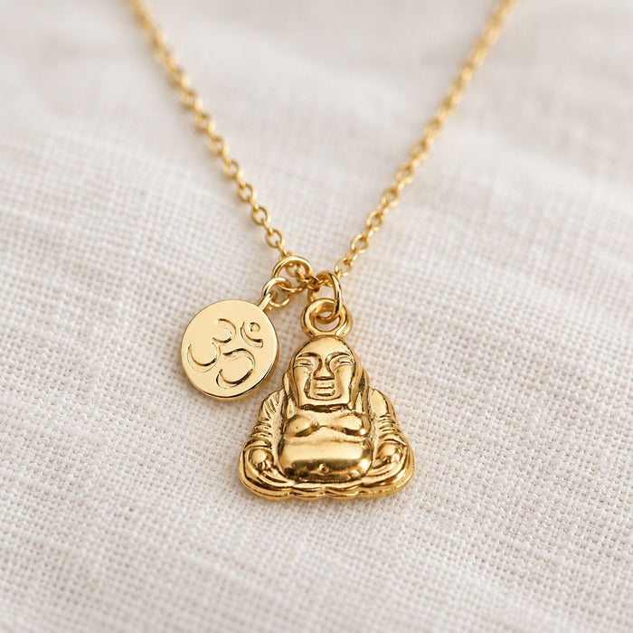 A gold-tone Buddha pendant necklace with a small Om symbol charm, displayed on a textured off-white linen fabric background.