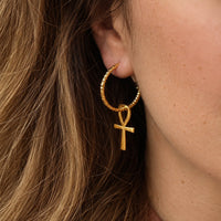 A close-up of a person's ear wearing a gold hoop earring with a dangling ankh symbol charm, shown against brown hair and skin.