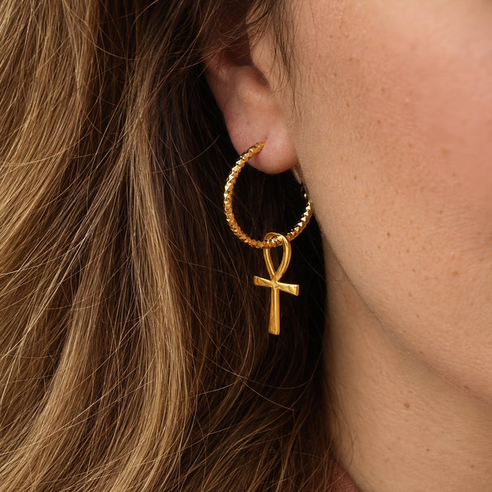 A close-up of a person's ear wearing a gold hoop earring with a dangling ankh symbol charm, shown against brown hair and skin.