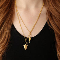 A close-up of a person wearing two layered gold necklaces over a black top. One necklace has a small sword-like pendant, and the other has a small starburst or flower-like pendant.
