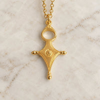 A gold pendant necklace with a stylized cross-like design and a small circular textured detail in the center, resting on a light marble surface.