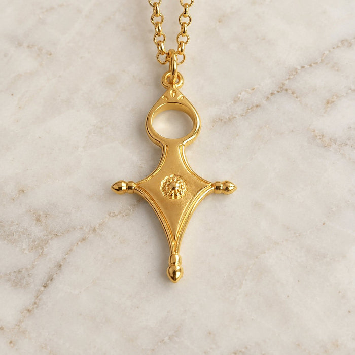A gold pendant necklace with a stylized cross-like design and a small circular textured detail in the center, resting on a light marble surface.